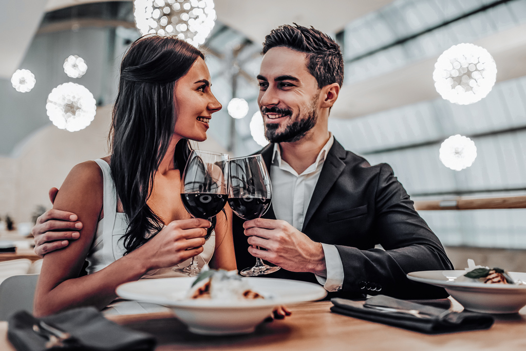 Beautiful loving couple is spending time together in modern restaurant. Attractive young woman in dress and handsome man in suit are having romantic dinner. Celebrating Saint Valentine's Day.